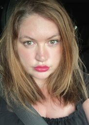 Close-up portrait of a person with light brown hair and hazel eyes, bright pink lipstick and a butterfly necklace, seated in a sunlit car with a seatbelt across the chest.