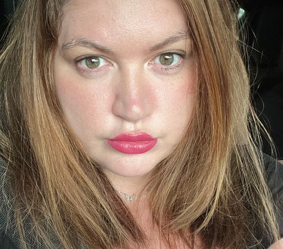 Close-up portrait of a person with light brown hair and hazel eyes, bright pink lipstick and a butterfly necklace, seated in a sunlit car with a seatbelt across the chest.