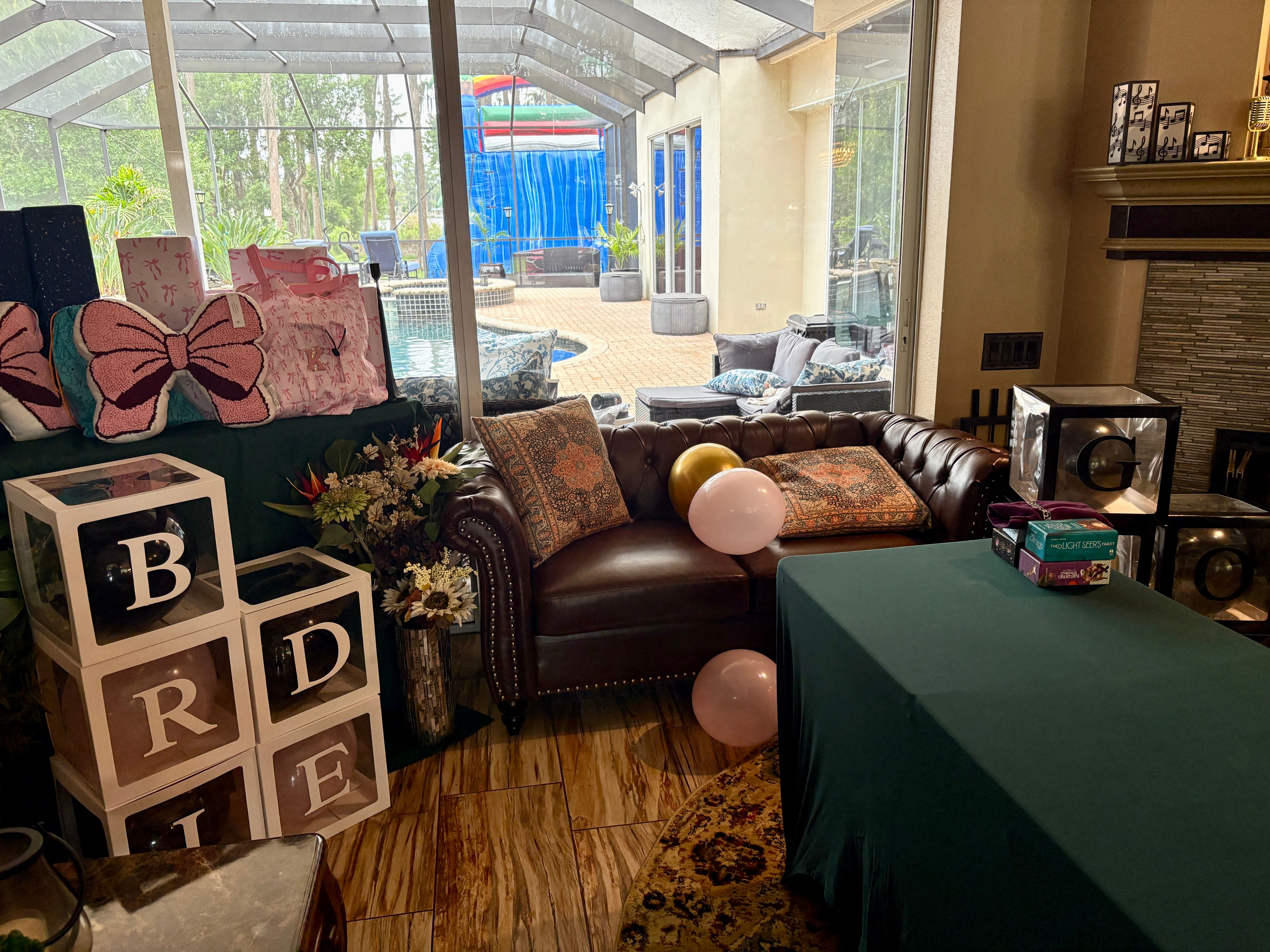 Living room party setup by screened pool patio: brown leather tufted sofa with patterned pillows, pink and gold balloons, stacked clear gift boxes and pink bow pillows, green-clothed table and backyard pool view.