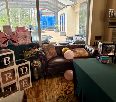 Living room party setup by screened pool patio: brown leather tufted sofa with patterned pillows, pink and gold balloons, stacked clear gift boxes and pink bow pillows, green-clothed table and backyard pool view.