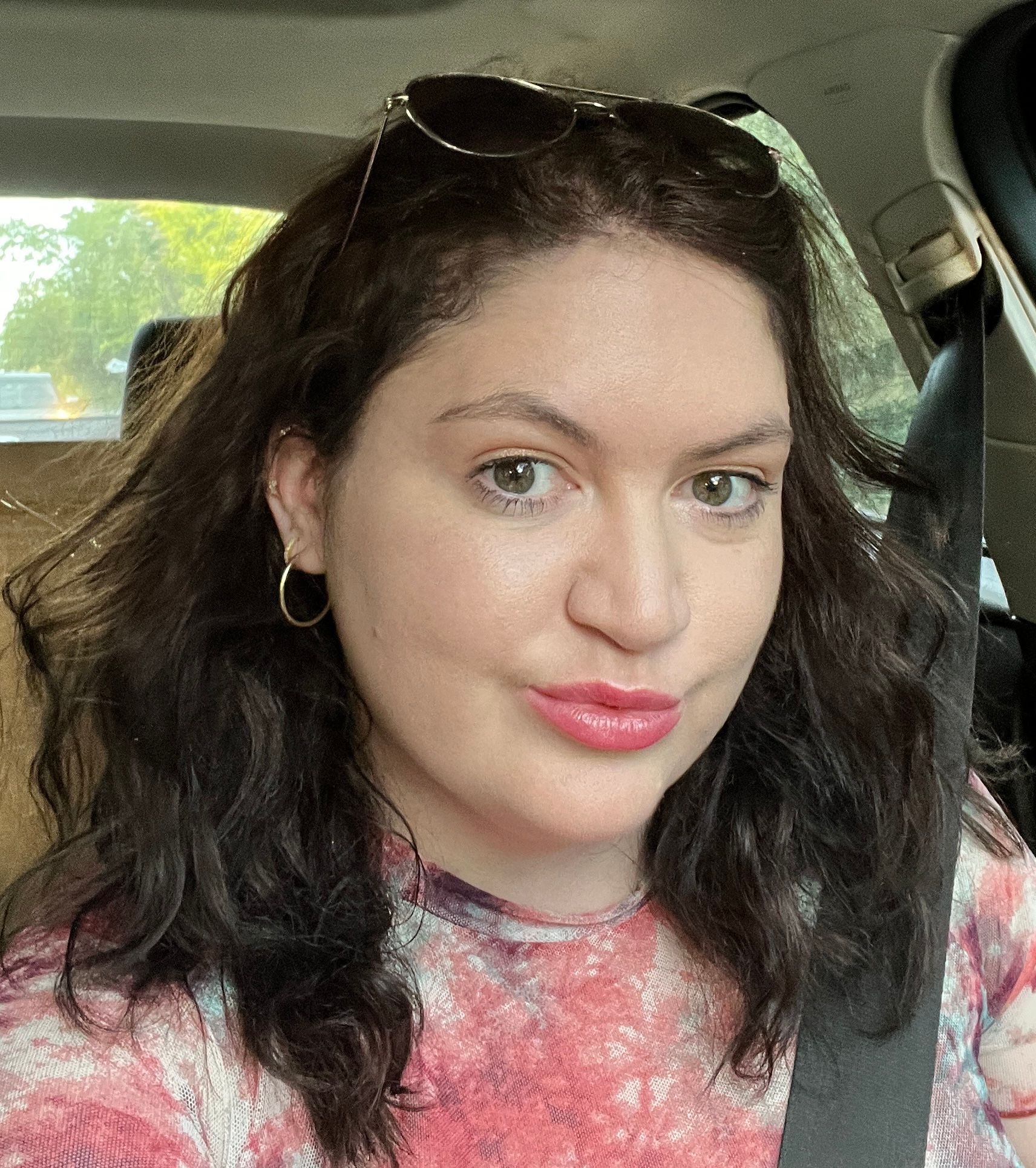 Close-up car selfie of a woman with wavy dark hair, green eyes, hoop earrings and sunglasses on her head, wearing a pink tie-dye top and a seatbelt