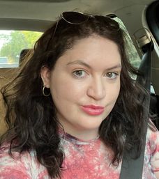 Close-up car selfie of a woman with wavy dark hair, green eyes, hoop earrings and sunglasses on her head, wearing a pink tie-dye top and a seatbelt