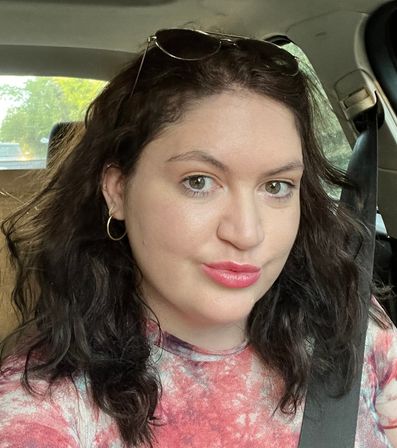 Close-up car selfie of a woman with wavy dark hair, green eyes, hoop earrings and sunglasses on her head, wearing a pink tie-dye top and a seatbelt