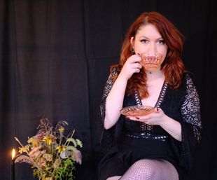 Red-haired woman in a black lace dress sips from a cut-glass teacup in a moody studio portrait with candlelight and a dried wildflower bouquet.