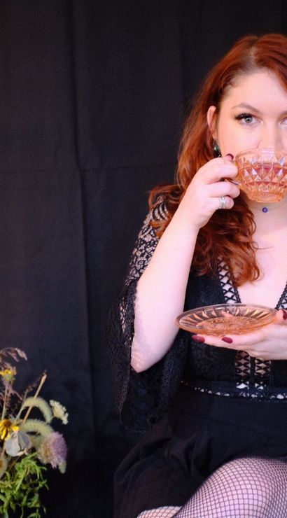 Red-haired woman in a black lace dress sips from a cut-glass teacup in a moody studio portrait with candlelight and a dried wildflower bouquet.