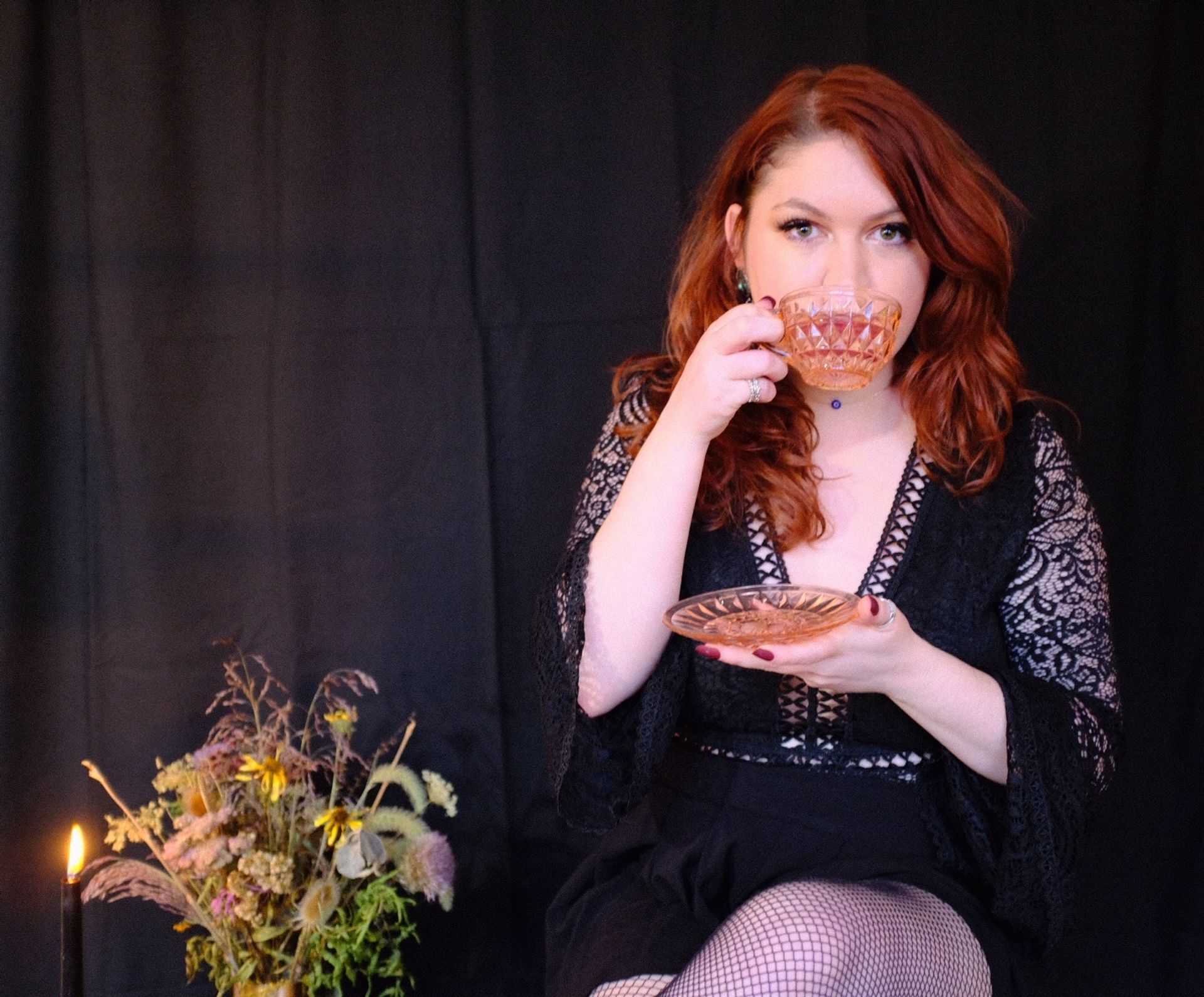 Red-haired woman in a black lace dress sips from a cut-glass teacup in a moody studio portrait with candlelight and a dried wildflower bouquet.