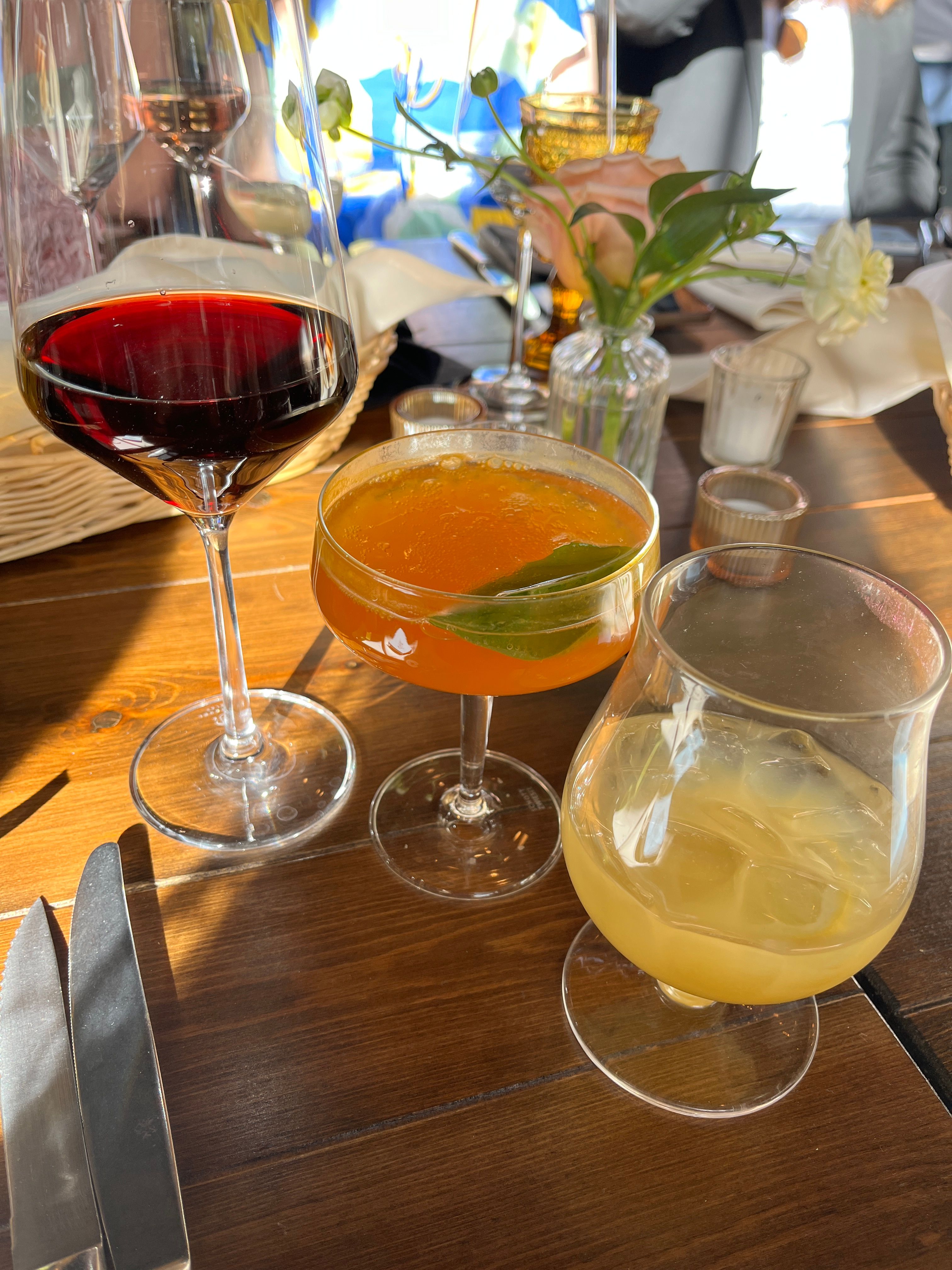 Sunlit restaurant table on warm wood with three drinks: a glass of red wine, an orange coupe cocktail garnished with a green leaf, and a pale iced cocktail, plus cutlery and a small flower vase