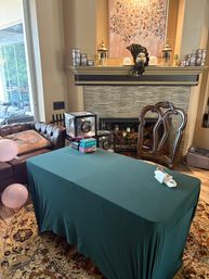 Cozy living room party setup with a green-clothed rectangular table in front of a tiled fireplace topped by a decorative mask and lanterns, a carved wooden chair, leather sofa, pink balloons and gift boxes nearby.