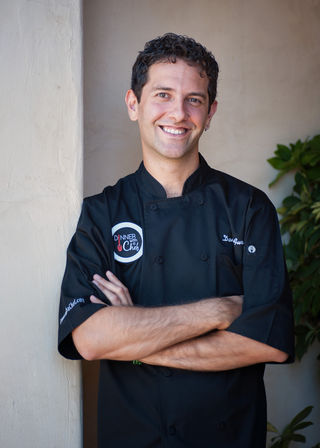 Friendly smiling chef in a black coat with arms crossed, leaning against a light stucco wall next to green plants — outdoor restaurant/patio portrait.