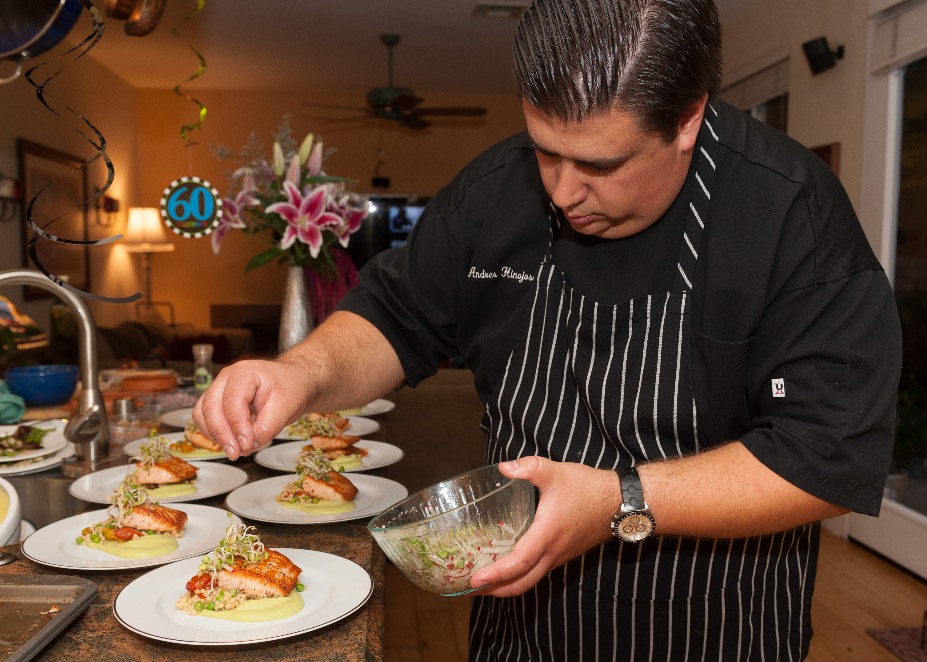Chef in striped apron plating seared salmon with microgreens and pea purée on white plates in a home kitchen, preparing multiple dishes for a 60th birthday celebration.