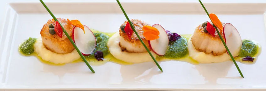 Three seared scallops on a white rectangular plate, each atop creamy purée with green herb sauce, radish slices, edible flowers and chive garnish — gourmet seafood appetizer, fine-dining restaurant plating