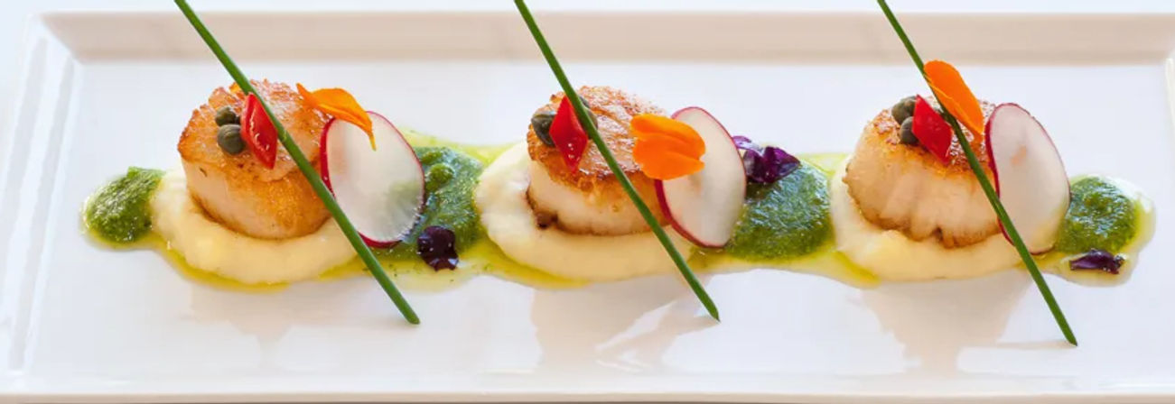 Three seared scallops on a white rectangular plate, each atop creamy purée with green herb sauce, radish slices, edible flowers and chive garnish — gourmet seafood appetizer, fine-dining restaurant plating