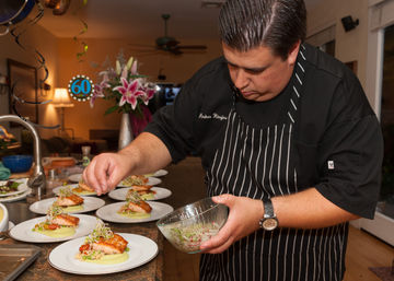 Chef in striped apron plating seared salmon on creamy puree with peas and sprouts on multiple white plates in a home kitchen, preparing dishes for a 60th birthday celebration.