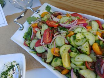 Bright summer cucumber and cherry tomato salad with red onion, arugula and parsley in a white serving dish on a rustic burlap-covered table