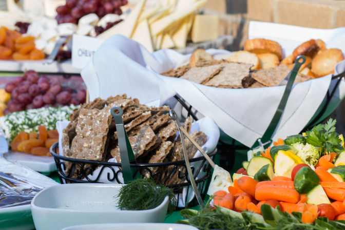Vibrant appetizer buffet/grazing table with baskets of seeded crispbreads and toasted bread, colorful vegetable platter of carrots and zucchini, bowls of dips, grapes and dried apricots