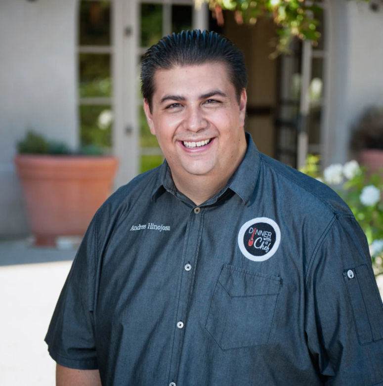 Smiling chef portrait on a sunny patio wearing a gray short-sleeve chef shirt with embroidered name and round logo patch, potted plants and glass doors in the background.