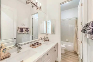 Bright modern residential bathroom with white double-sink vanity, large wall mirror, chrome faucets, woven tray accessories, and tub-shower with beige curtain and wood-look flooring.