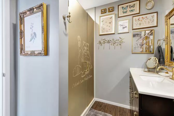 Residential bathroom with gold-accented etched shower door showing a playful animal sketch, white vanity with gold mirror and fixtures, gallery wall of botanical and butterfly prints, and wood-look flooring — stylish modern home bath.
