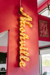 Bold vertical yellow marquee sign studded with glowing vintage bulbs mounted on a hot-pink interior wall, with a framed playing-card poster and windowed seating area visible in the background.