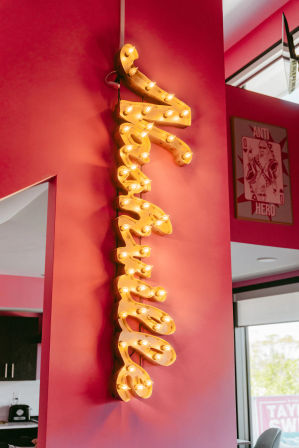 Bold vertical yellow marquee sign studded with glowing vintage bulbs mounted on a hot-pink interior wall, with a framed playing-card poster and windowed seating area visible in the background.