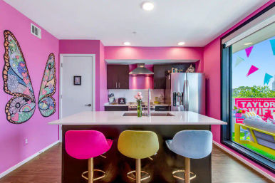 Vibrant pink modern apartment kitchen with a white island and sink, three colorful bar stools (pink, yellow, blue), whimsical butterfly wall mural, stainless steel appliances, and a sliding glass door to a balcony with pennant flags and a fan sign.