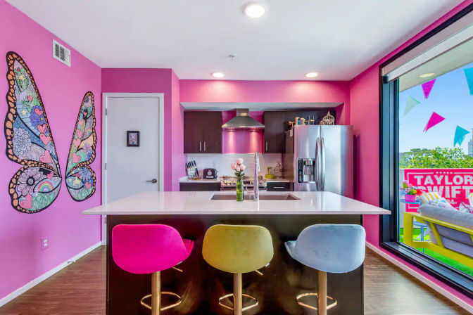 Vibrant pink modern apartment kitchen with a white island and sink, three colorful bar stools (pink, yellow, blue), whimsical butterfly wall mural, stainless steel appliances, and a sliding glass door to a balcony with pennant flags and a fan sign.