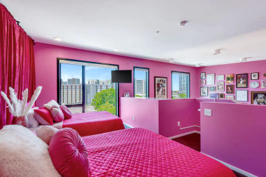 Vibrant pink loft bedroom with two twin beds in quilted hot-pink covers and round pillows, large windows showing a downtown city skyline and trees, gallery wall of framed art, wall-mounted TV and matching pink curtains.