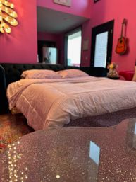 Playful bright pink bedroom interior with queen bed in pale pink bedding, glitter-speckled glass coffee table in foreground, dark tufted headboard, wall-mounted acoustic guitar and bold magenta walls.