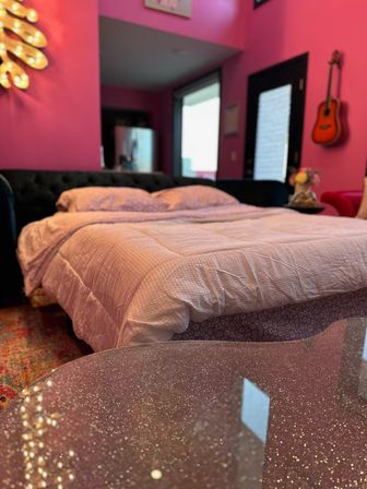 Playful bright pink bedroom interior with queen bed in pale pink bedding, glitter-speckled glass coffee table in foreground, dark tufted headboard, wall-mounted acoustic guitar and bold magenta walls.