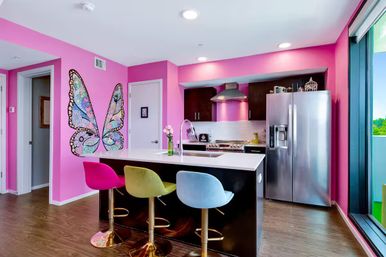 Bright pink modern apartment kitchen with a large butterfly mural, white island and sink, stainless-steel refrigerator, and three colorful bar stools (pink, green, blue) beside a sliding glass door overlooking greenery.
