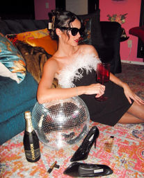Person in a black feather-trim dress and sunglasses lounging on a colorful rug, holding a red wine glass beside a disco ball, champagne bottle, black patent heels and perfume in a retro-glam living room with velvet sofa and bright pillows.