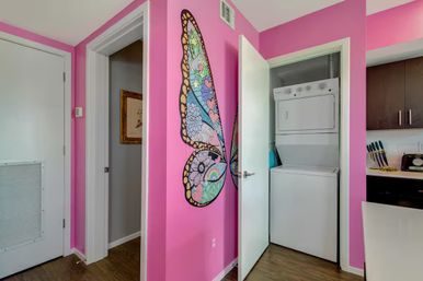 Apartment hallway with hot-pink accent wall and playful butterfly-wing mural, open laundry closet with stacked washer-dryer, and adjacent compact kitchen with dark cabinets.