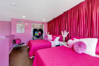 Vibrant pink bedroom with two twin beds dressed in quilted fuchsia bedding, round and heart-shaped pillows, large hot-pink floor-to-ceiling curtains, wood floors, and a sliding frosted glass door featuring a black heart decal and colorful window-like design.