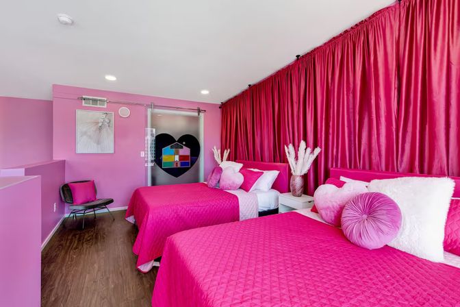 Vibrant pink bedroom with two twin beds dressed in quilted fuchsia bedding, round and heart-shaped pillows, large hot-pink floor-to-ceiling curtains, wood floors, and a sliding frosted glass door featuring a black heart decal and colorful window-like design.