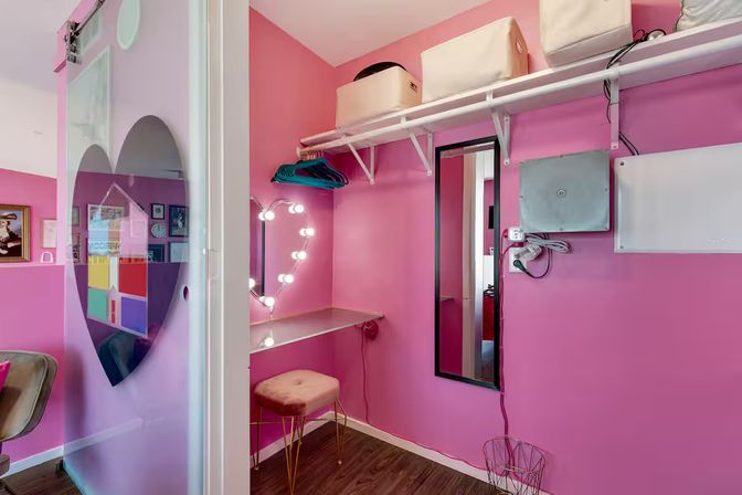 Bright pink dressing nook with a lighted heart-shaped mirror, velvet stool, floating vanity shelf, hanging rack and frosted heart sliding door
