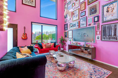 Vibrant pink urban apartment living room with floor-to-ceiling windows and city view, eclectic gallery wall, velvet blue and red sofas, wall guitar, and mirrored disco-ball coffee table.