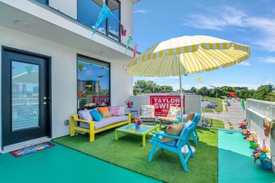 Sunny urban balcony patio with yellow-striped umbrella, vibrant blue and yellow seating, green turf, colorful bunting and a red campaign-style sign overlooking city streets.