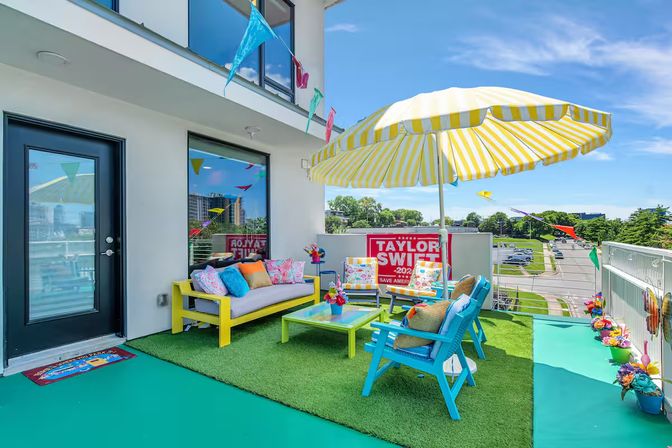 Sunny urban balcony patio with yellow-striped umbrella, vibrant blue and yellow seating, green turf, colorful bunting and a red campaign-style sign overlooking city streets.
