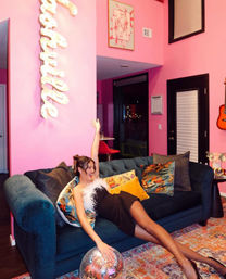 Woman in a black feather-trimmed dress lounges on a teal velvet sofa in a vibrant pink living room, reaching toward a disco ball on a colorful rug — playful decor includes patterned pillows, a wall-mounted acoustic guitar and illuminated script sign.
