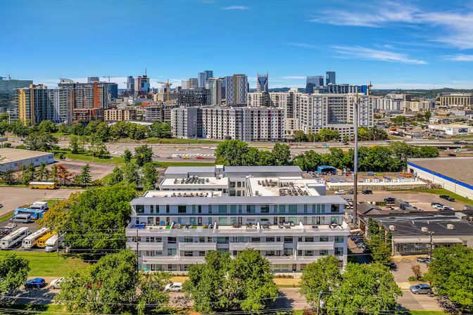 Aerial view of a modern mid-rise apartment complex with balconies and rooftop equipment, tree-lined street and parking in the foreground, a multi-lane highway and dense downtown skyline under a bright blue sky.