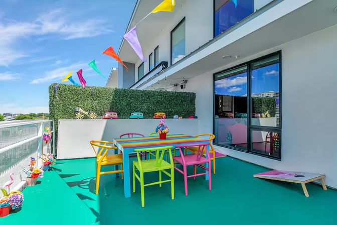 Bright urban rooftop balcony with rainbow-striped dining table and multicolored chairs on a turquoise floor, colorful pennant flags, faux hedge with toy car decor, potted flowers and clear blue sky