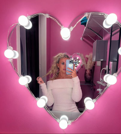 Mirror selfie in a pink room: woman in a white off-shoulder sweater posing in a heart-shaped vanity mirror lined with round bulbs, phone partially covering her face, shelf with vase and pampas grass in the background.