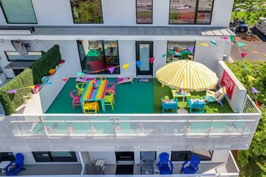 Colorful rooftop apartment balcony on a modern white building with a rainbow dining table, yellow-striped umbrella, bunting flags and bright lounge seating.