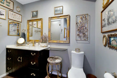 Chic vintage-inspired powder room with dark wood vanity, white countertop, gold faucet and ornate gold-framed mirrors, gallery wall art, potted lavender, tufted round stool, and white toilet on soft gray walls.