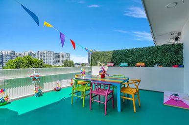 Colorful rooftop balcony with a rainbow-striped dining table and mismatched bright chairs on a turquoise floor, festive pennant flags overhead, faux hedge wall lined with toy camper vans, potted flowers and a cornhole board, city high-rise skyline beyond.