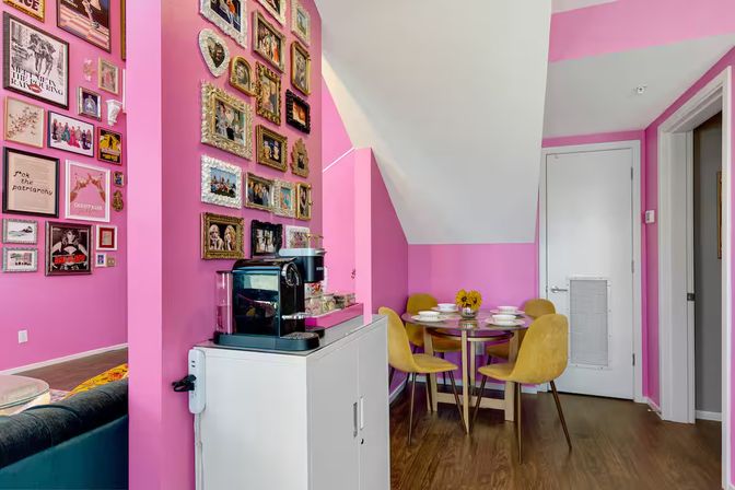 Vibrant pink apartment dining nook with a gallery wall of ornate frames, small round table set for four with mustard-yellow chairs, adjacent white cabinet coffee station and hardwood floors.