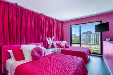 Vibrant pink twin bedroom in a downtown apartment with quilted bedding, round accent pillows, floor-to-ceiling curtains and a large window showing an urban skyline view.