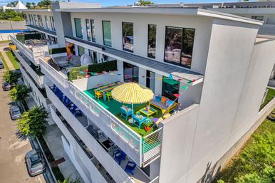 Top-floor balcony rooftop terrace on a modern apartment building with a yellow-striped umbrella, colorful chairs and tables, pennant flags, green turf and cozy seating overlooking the street