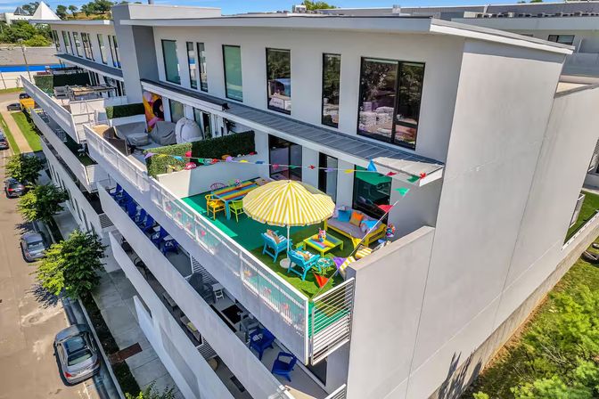 Top-floor balcony rooftop terrace on a modern apartment building with a yellow-striped umbrella, colorful chairs and tables, pennant flags, green turf and cozy seating overlooking the street