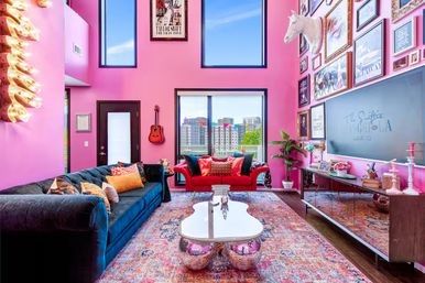 Vibrant pink urban loft living room with double-height windows and city skyline view, navy velvet sofa and red loveseat with colorful throw pillows, patterned rug, mirrored guitar-shaped coffee table on disco-ball bases, wall-mounted guitar, eclectic framed gallery wall with a white unicorn head sculpture, plants and mirrored media console.